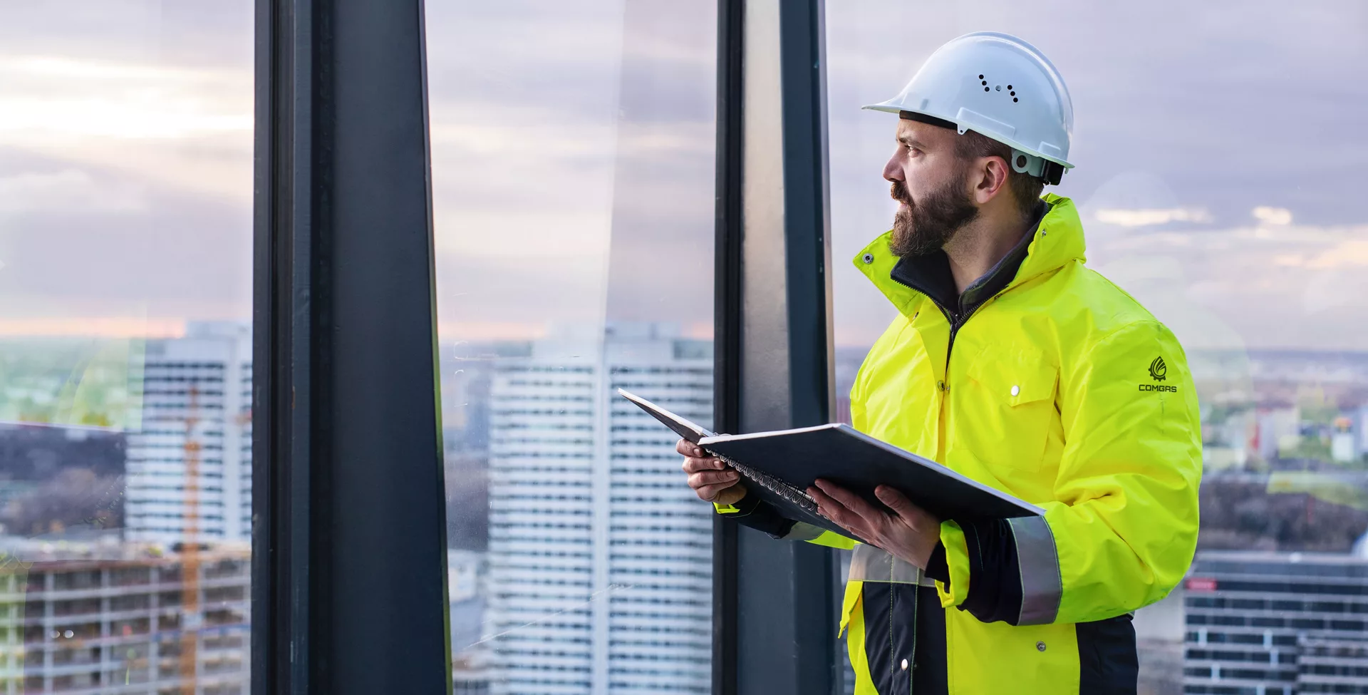 Engineer in ComGas hi-vis jacket and hard hat reviewing site documents beside a high-rise window