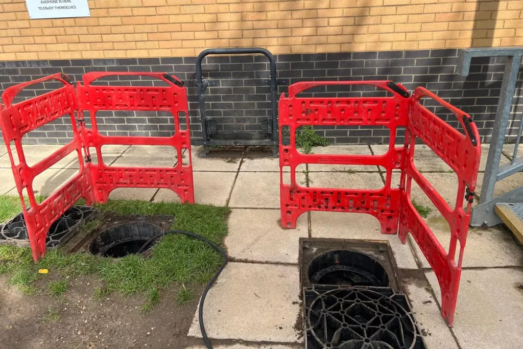Two open drainage inspection chambers beside a clubhouse wall, barriered off with red safety fencing, with a hose run between them