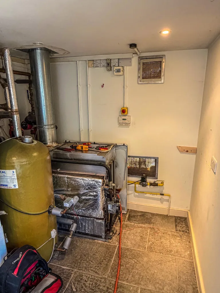 The original floor-standing boiler and hot water cylinder in the plant cupboard before replacement, with the boiler casing opened and its flue rising to the ceiling