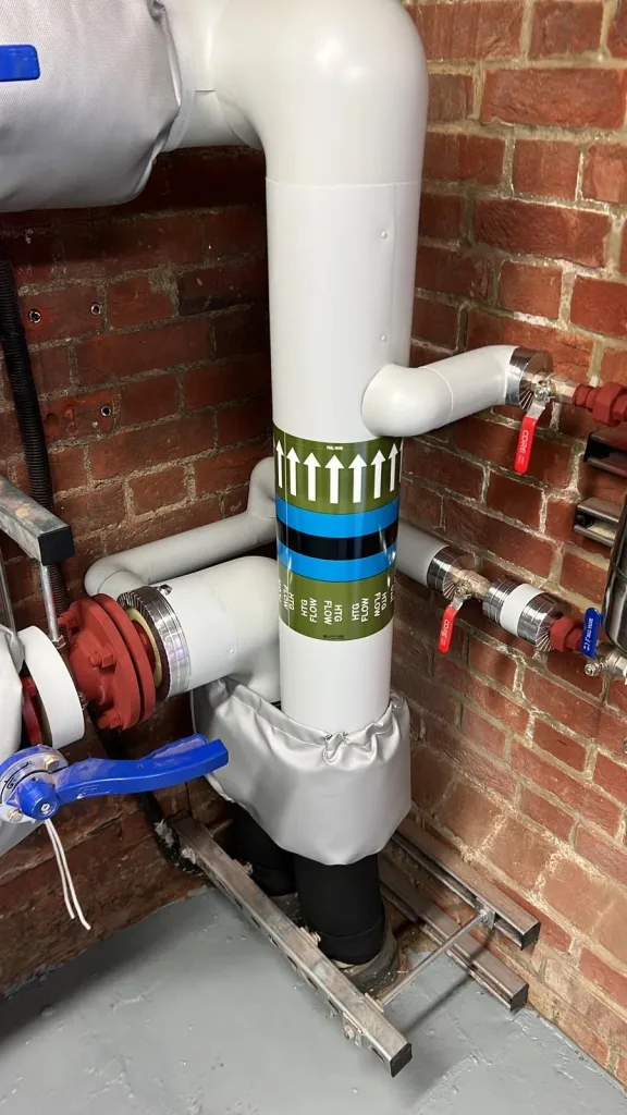 Vertical insulated heating pipe with green and blue HTG FLOW identification bands, lever valves and channel supports.