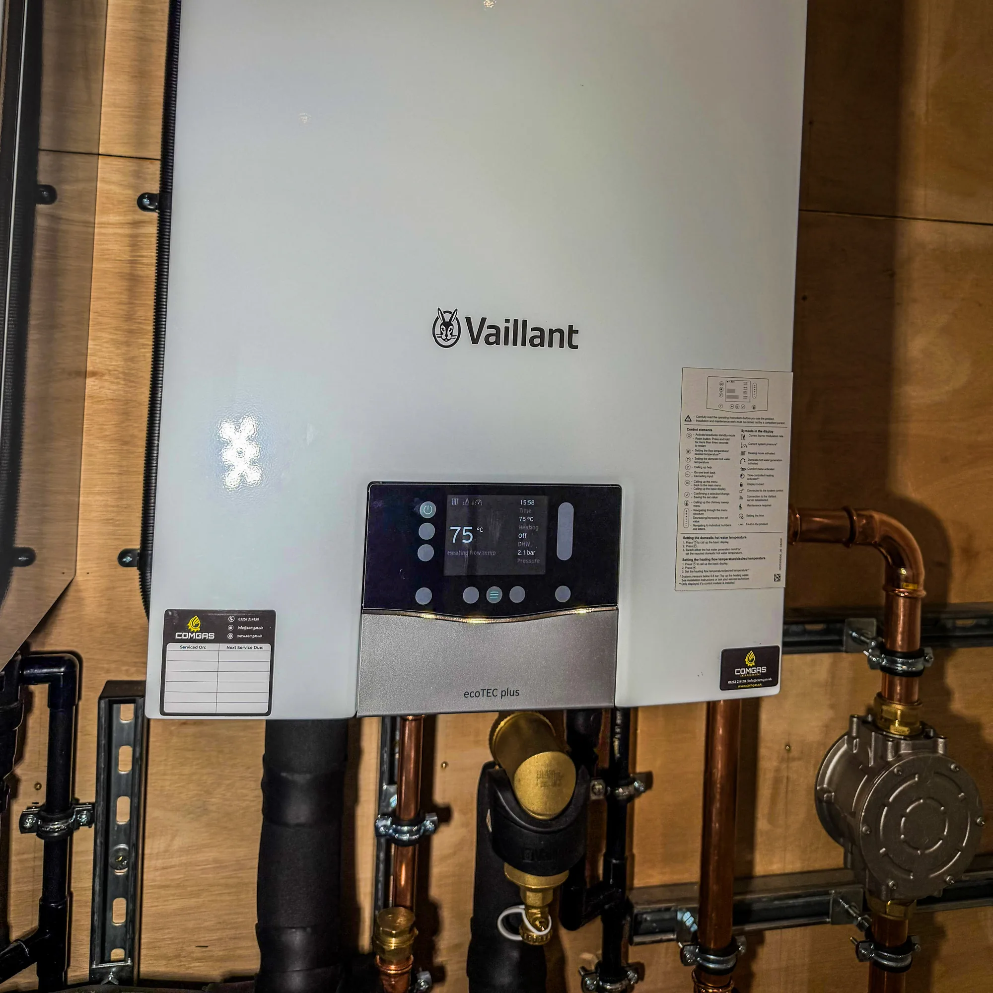 Close-up of a Vaillant ecoTEC plus boiler installed by ComGas, its display reading 75 degrees and 2.1 bar, with a ComGas service record sticker on the casing